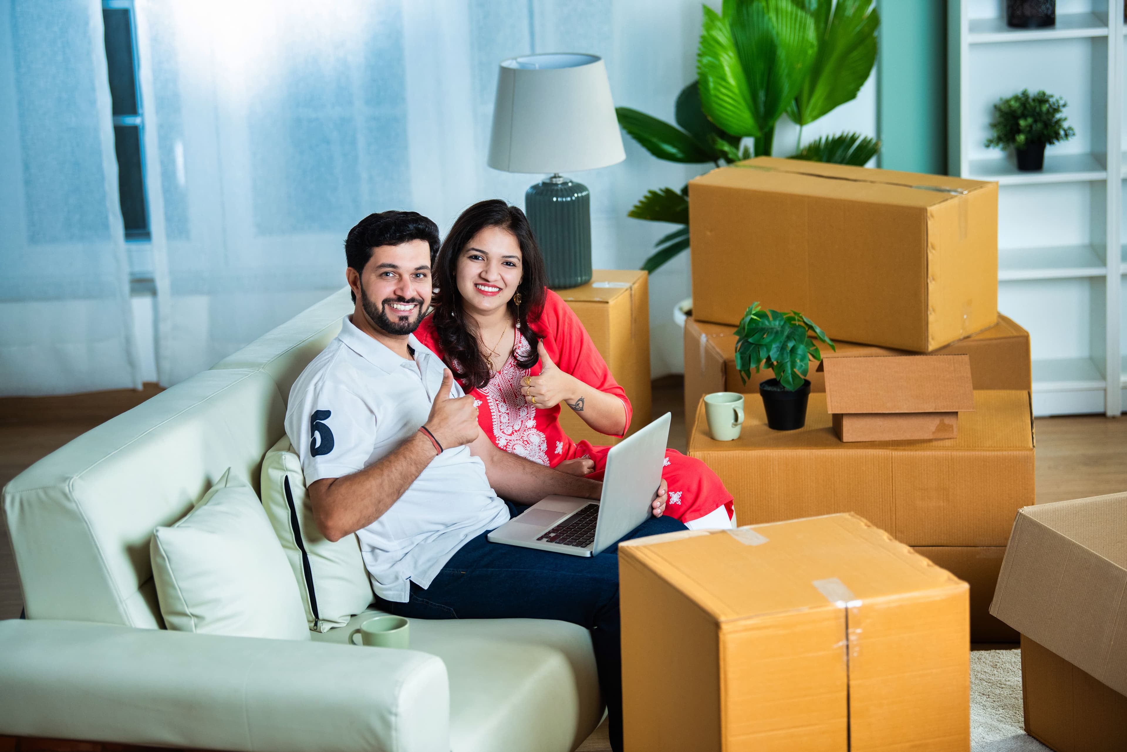 Young Indian couple using a laptop on a sofa amidst home shifting with moving boxes around